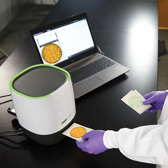 A laboratory technician wearing purple gloves uses the Neogen Petrifilm™ Plate Reader Advanced, placing prepared Petrifilm plates into the imaging device connected to a laptop displaying digital colony count results.